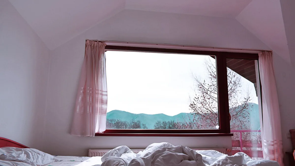 Dormitorio luminoso con la cama deshecha frente a una ventana con vistas a la montaña.