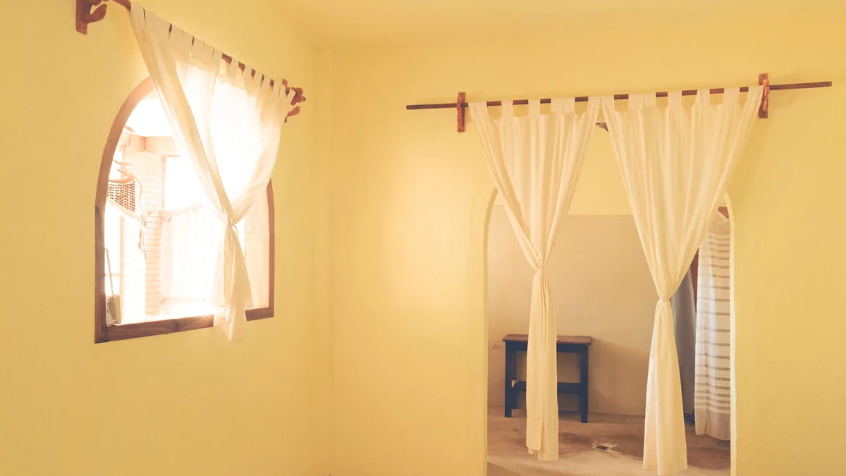 Visillos blancos colgados de una barra de madera fijada a la pared, uno recogido junto a una ventana en arco, en una habitación de paredes amarillas.